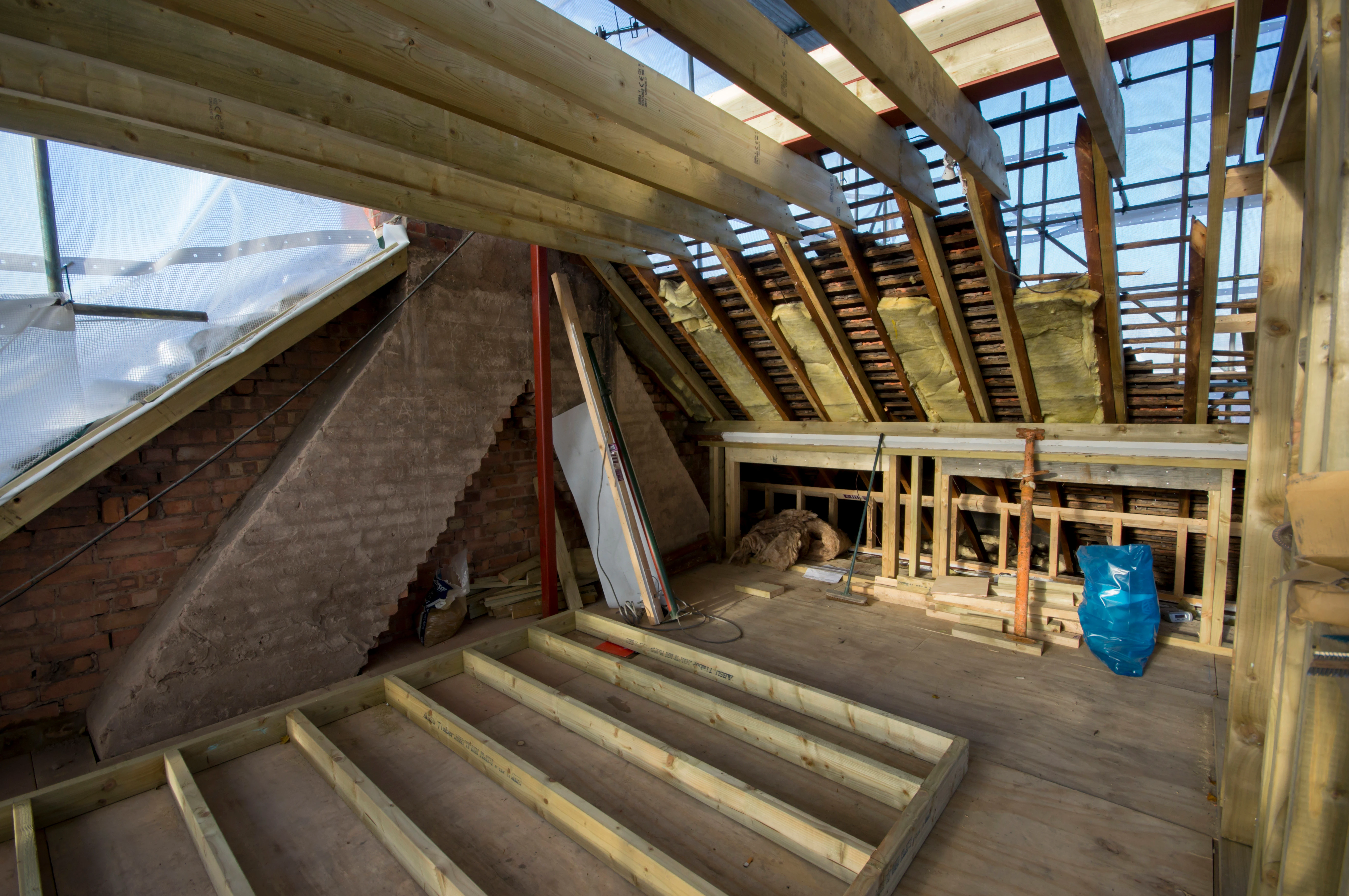 Interior of an under-construction attic showing wooden frames, exposed brick wall, and scaffolding for roof work.