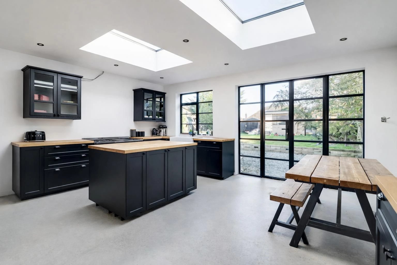 Modern kitchen with black cabinets, wooden countertops, skylights, and large windows overlooking a garden, featuring a wooden dining table.