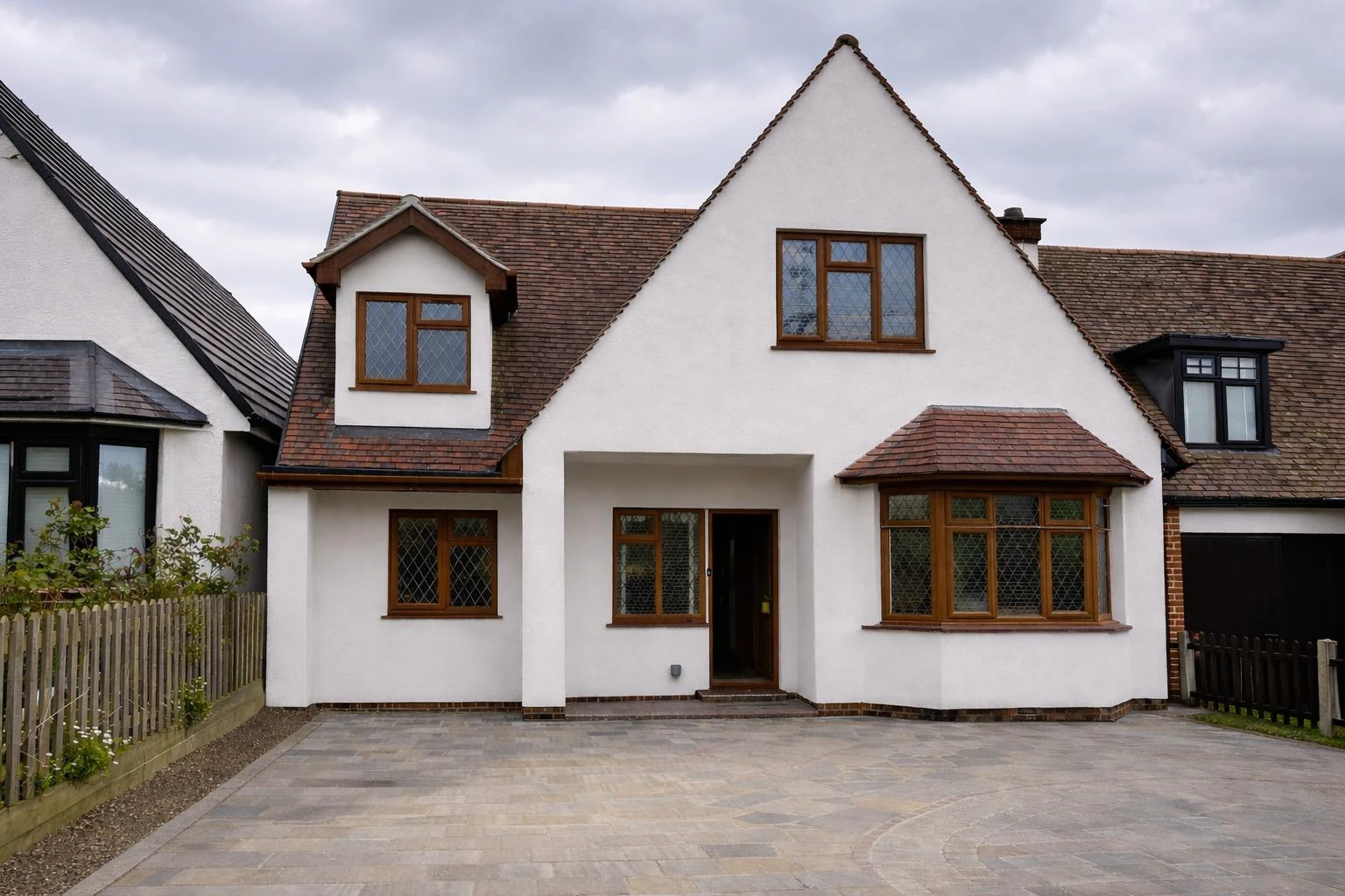 A white house with a gabled roof and brown window frames, surrounded by a paved driveway and leafy plants.