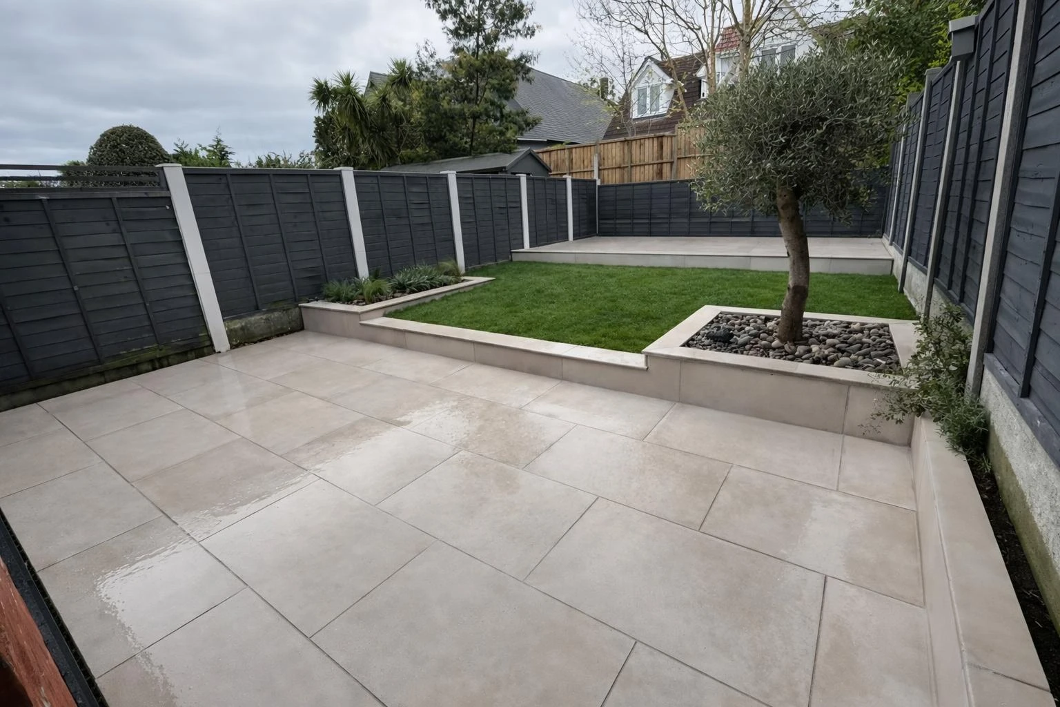 Modern backyard featuring gray paving slabs, a green lawn, and decorative stones, enclosed by dark wooden fencing.