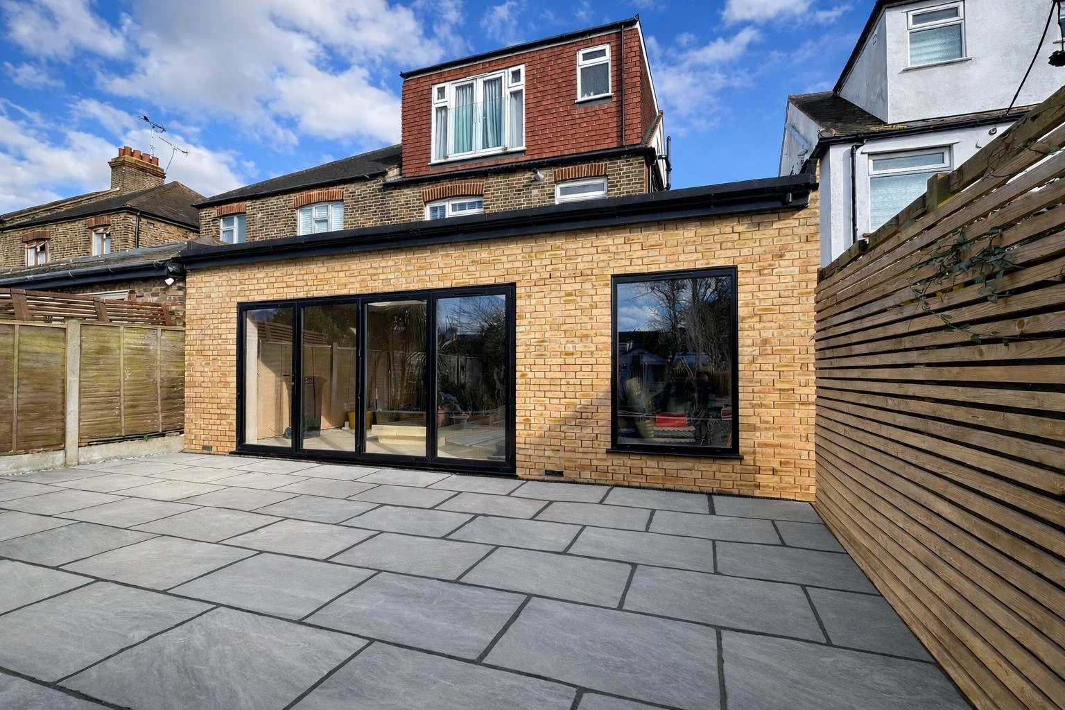 Modern brick house with large glass doors, surrounded by a spacious patio and wooden fencing, under a blue sky with clouds.