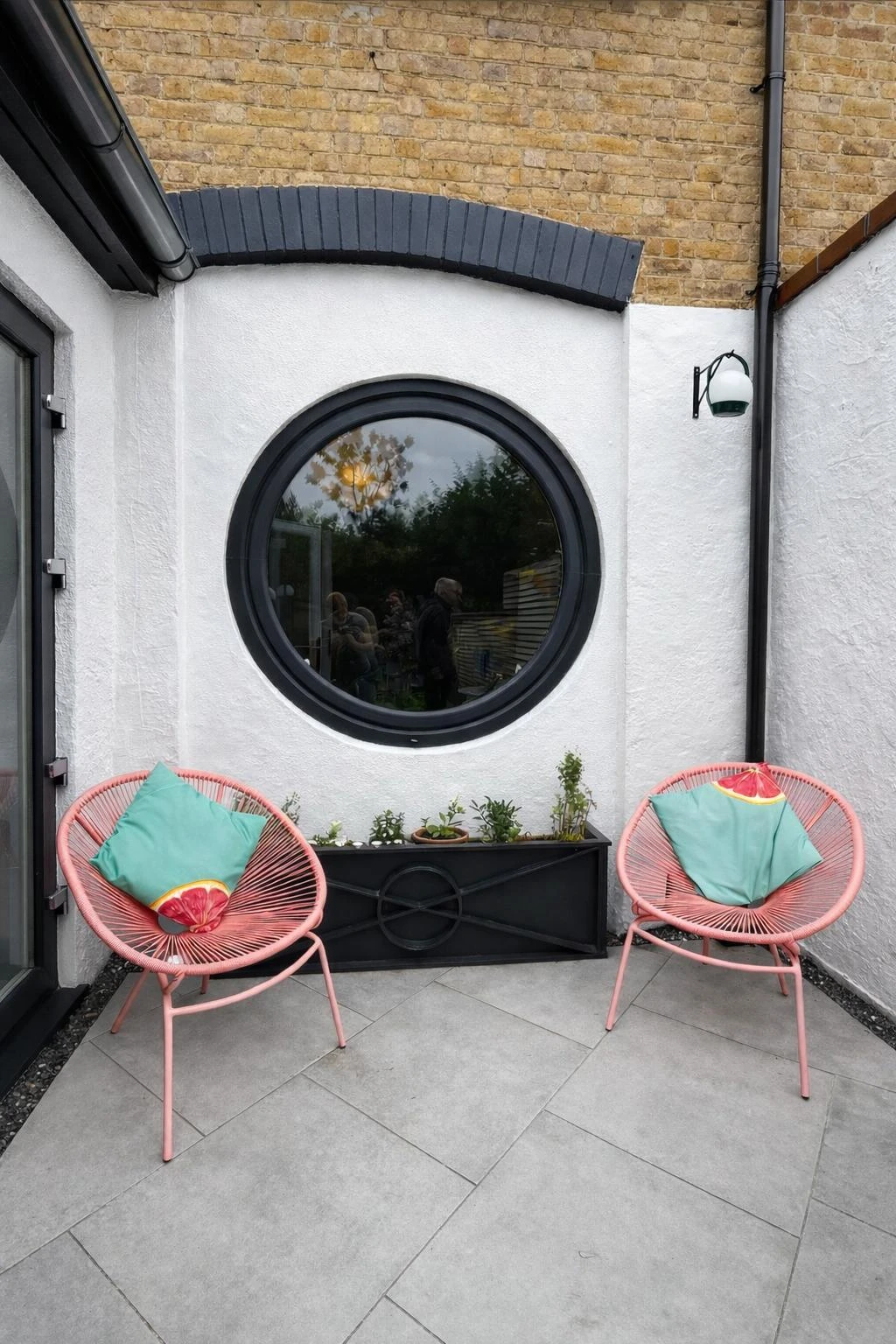 Cozy outdoor seating area featuring two pink chairs with colorful cushions and a round black window, surrounded by plants and gray tiles.