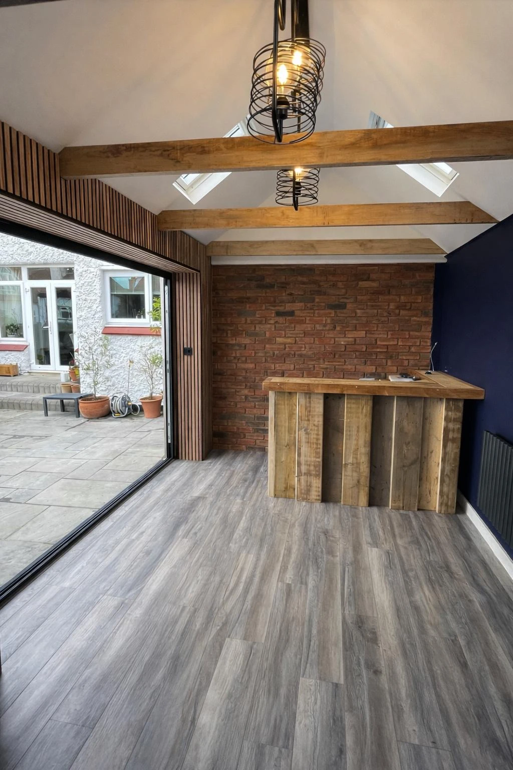 Modern interior space featuring wooden beams, a brick wall, and a wooden bar on a gray floor, with large glass doors opening to an outdoor area.