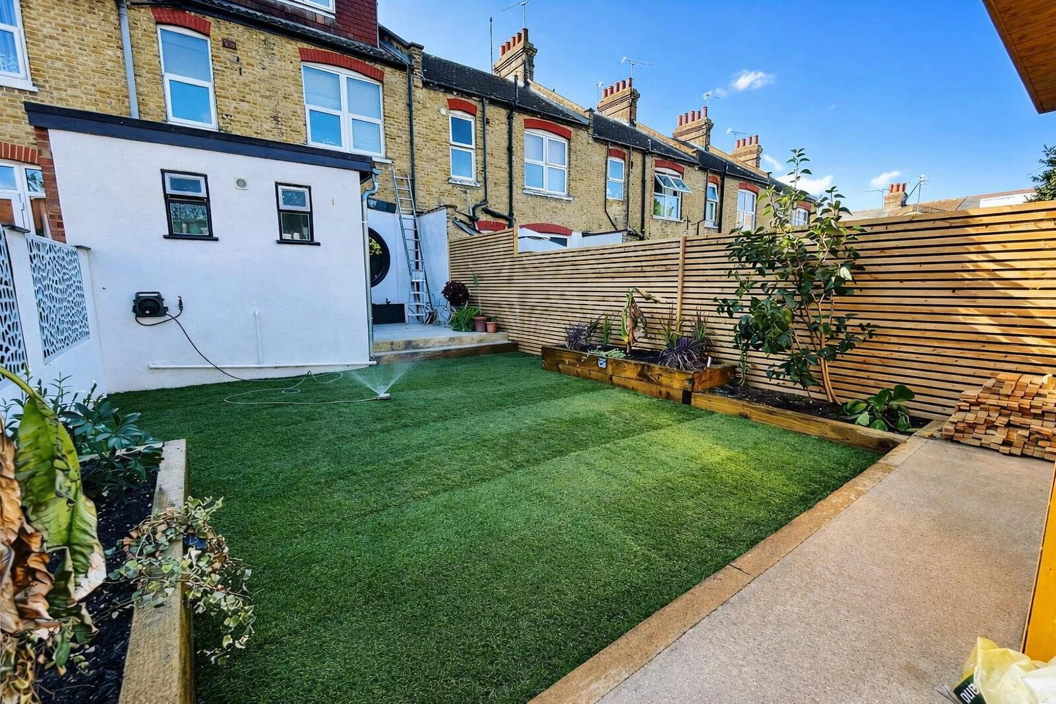 Lush green garden with artificial turf, wooden planters, and a fence, featuring a house in the background and a ladder leaning against the wall.
