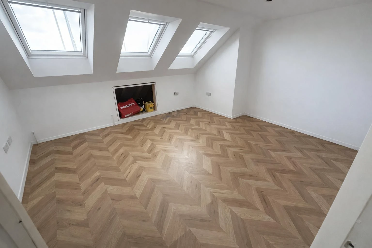 Bright attic room featuring angled skylights and herringbone wood flooring. A small storage area is visible in the wall.