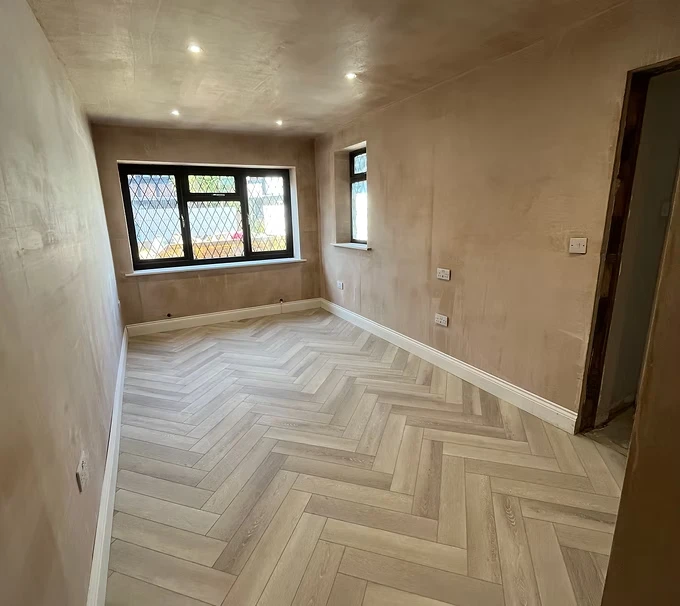 Empty room with light-colored herringbone flooring, beige walls, and multiple windows allowing natural light.